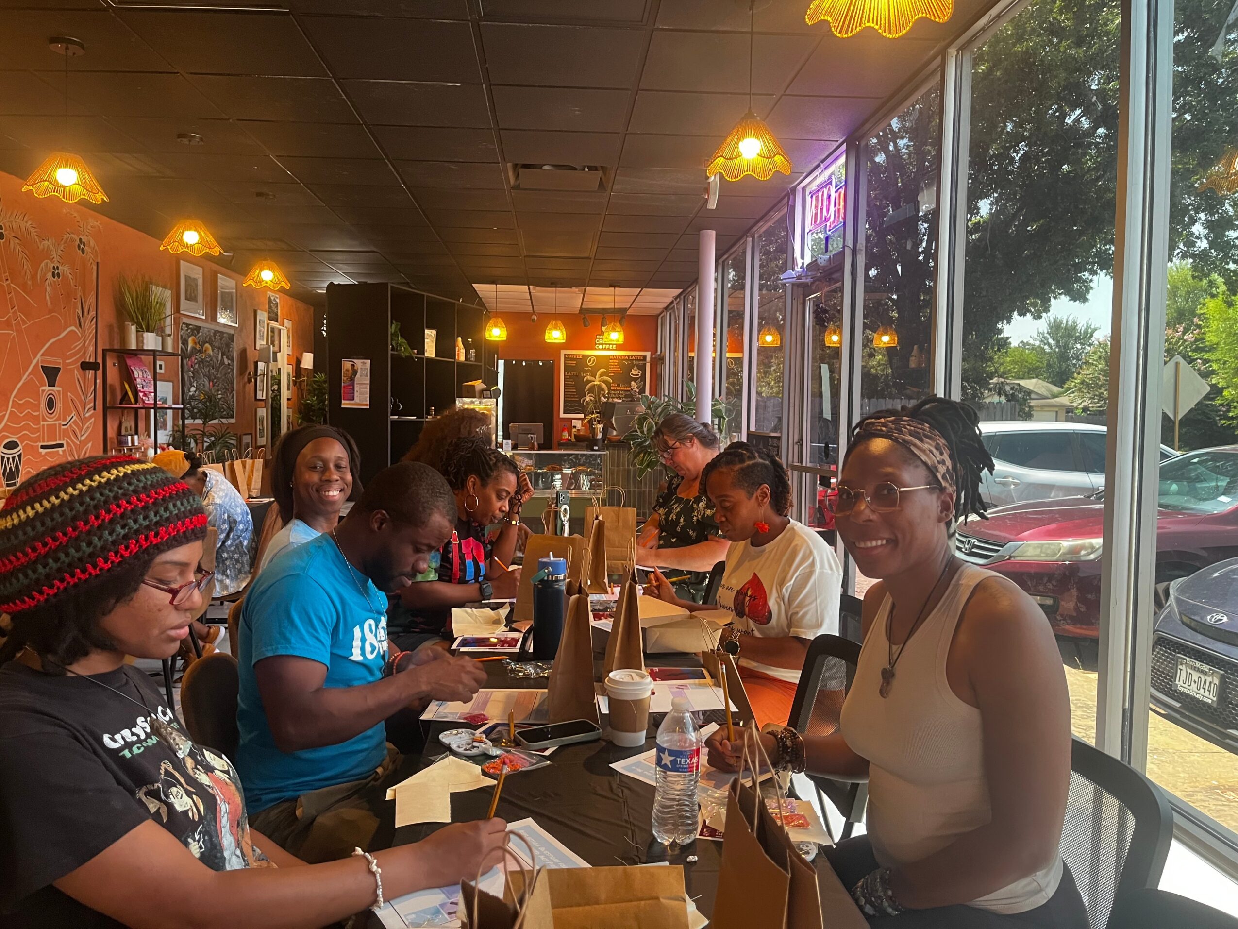 Group of people sitting at a long table inside a restaurant with large windows and warm lighting, enjoying food and drinks.