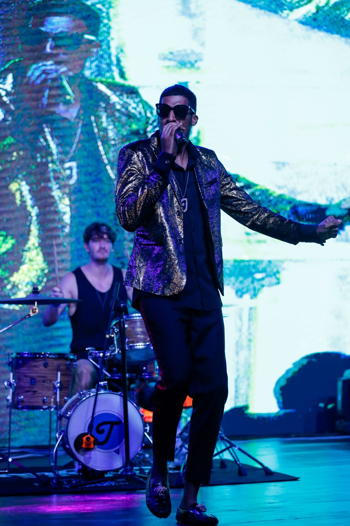Performer singing into microphone on stage with band, large screen behind displaying his image, colorful lighting, and musical instruments.