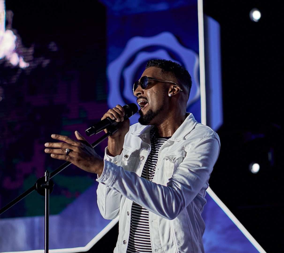 Male singer performing on stage with microphone, wearing sunglasses and a white jacket, background with blue and purple lighting.