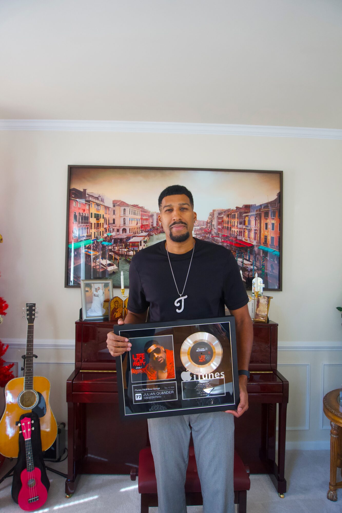 Man holding a framed award in a room with a piano, guitar, and colorful artwork on the wall behind him.