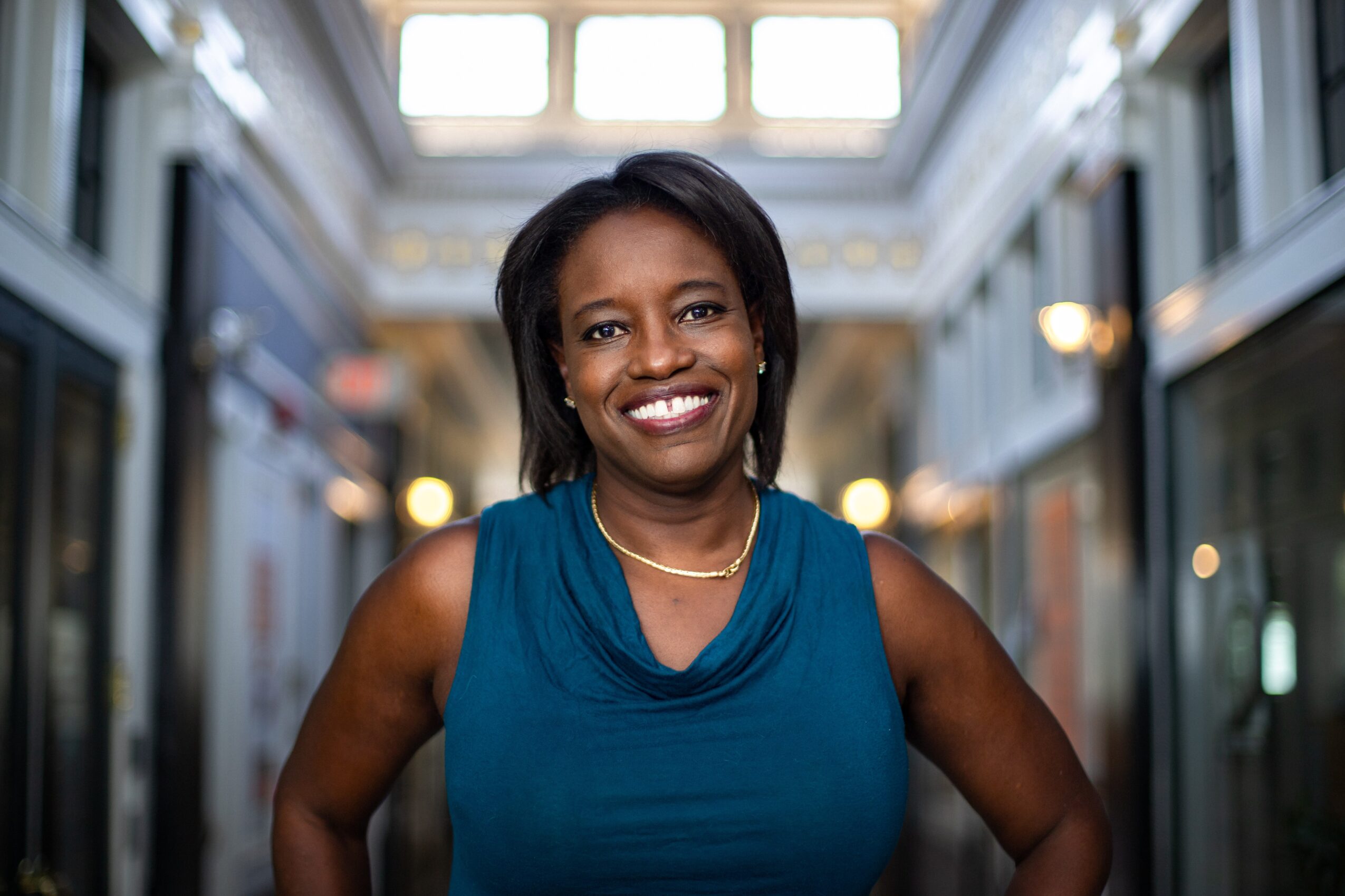 Smiling woman in a blue sleeveless top standing in a bright indoor shopping area with glass ceiling.