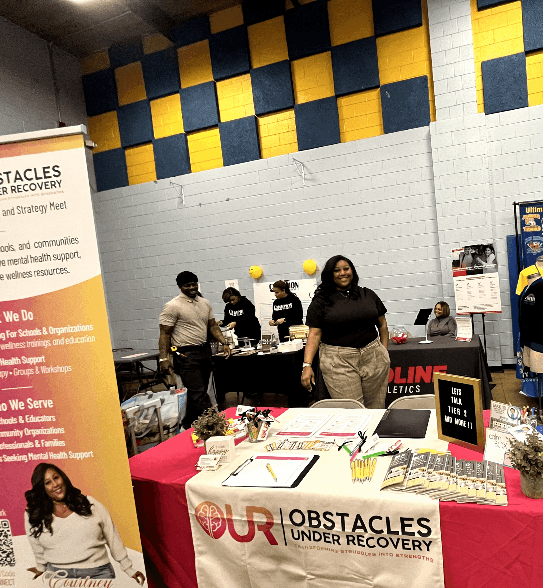 People at a table with a banner reading 'Obstacles Under Recovery' in a room with a colorful wall, flowers, and informational materials.