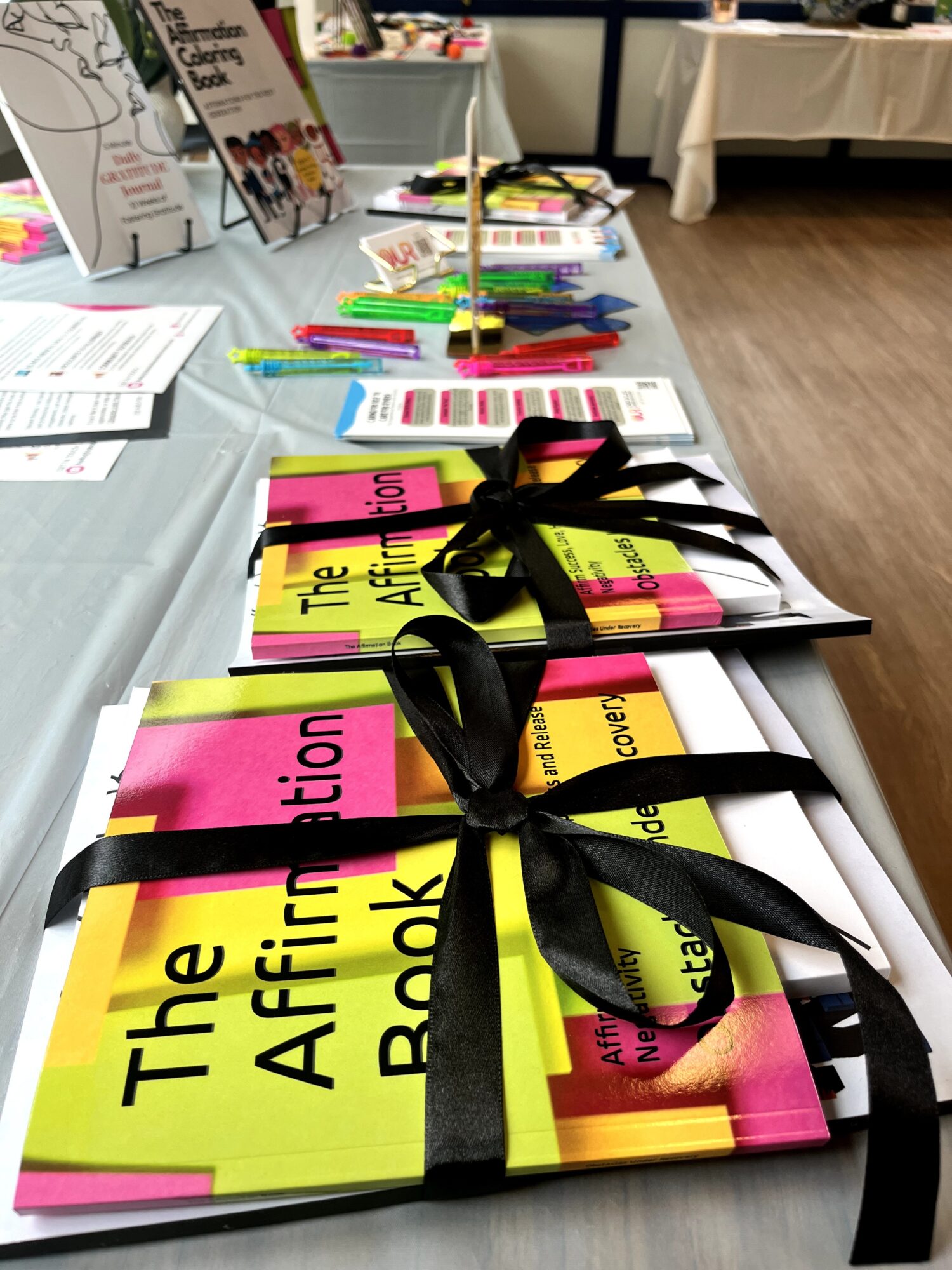 Table with colorful booklets tied with black ribbons, displayed in a row, with other materials and tables in background.