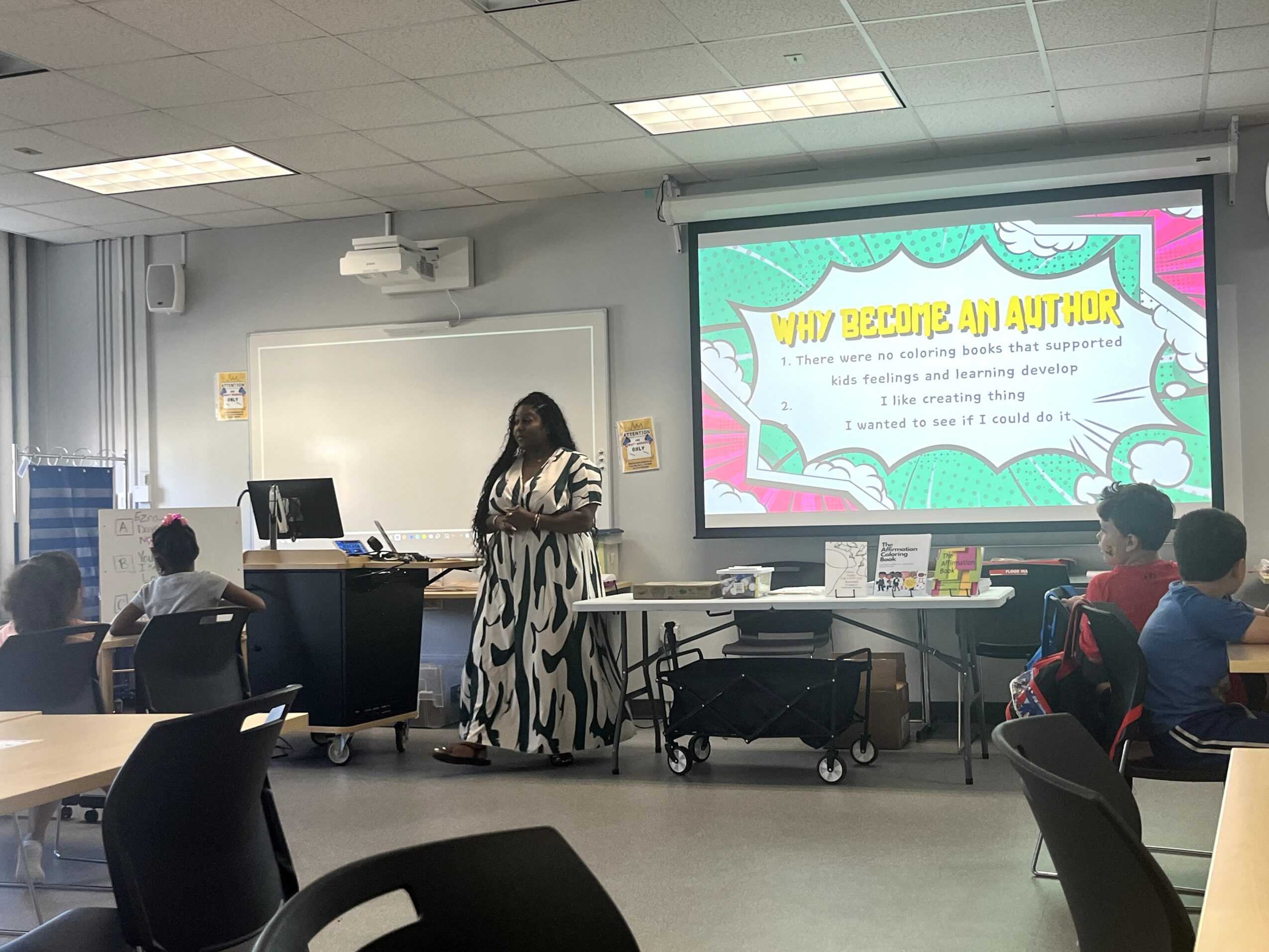 A woman stands at the front of a classroom giving a presentation to students seated at desks, with a colorful slide projected behind her.