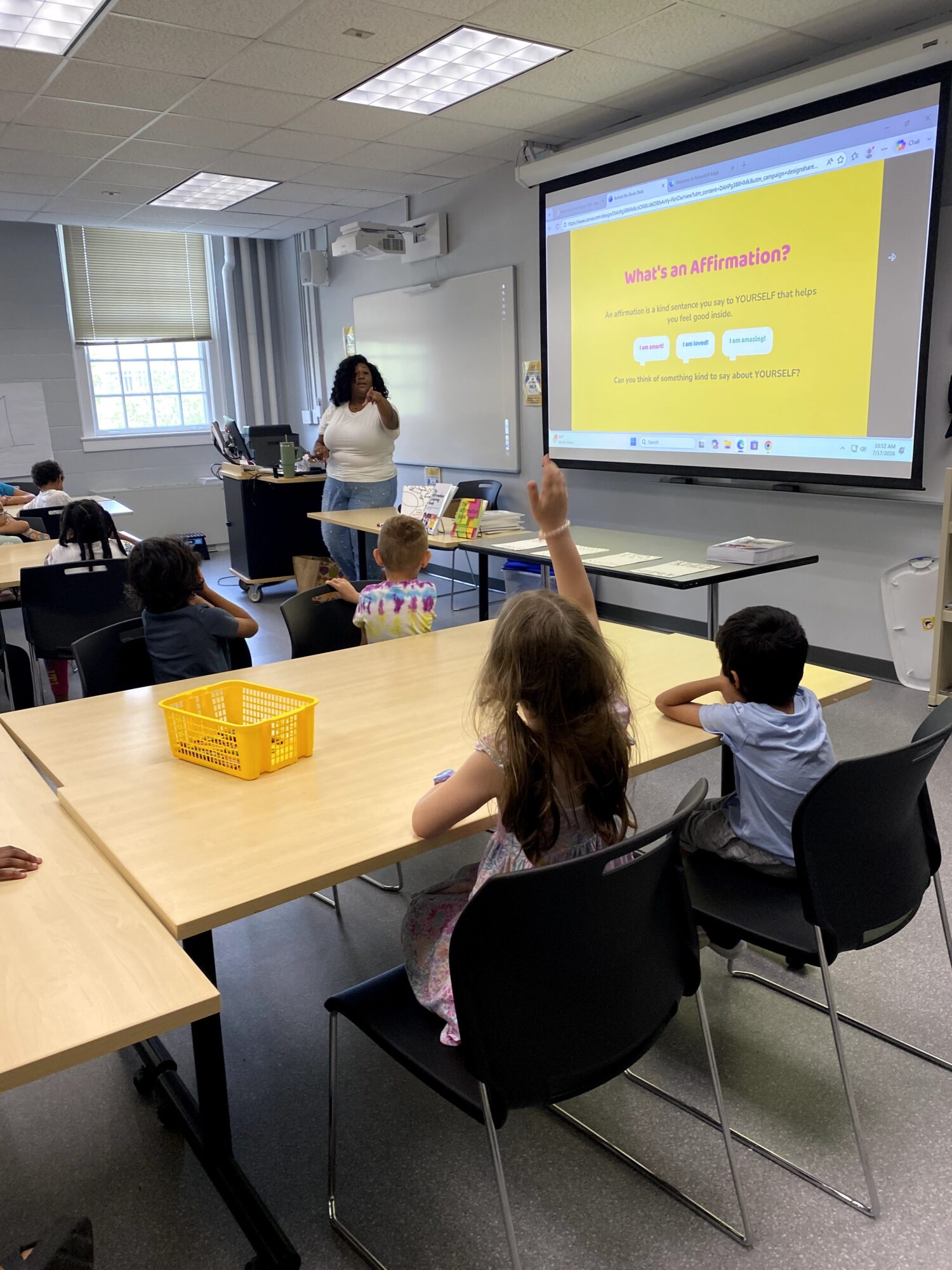 Classroom with children seated at tables, teacher at front, large screen displaying a yellow slide, some children raising hands.