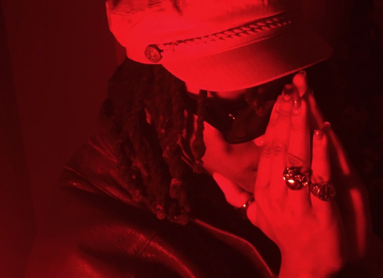 Person wearing a cap, sunglasses, and rings, with hands pressed together near face, in red lighting.