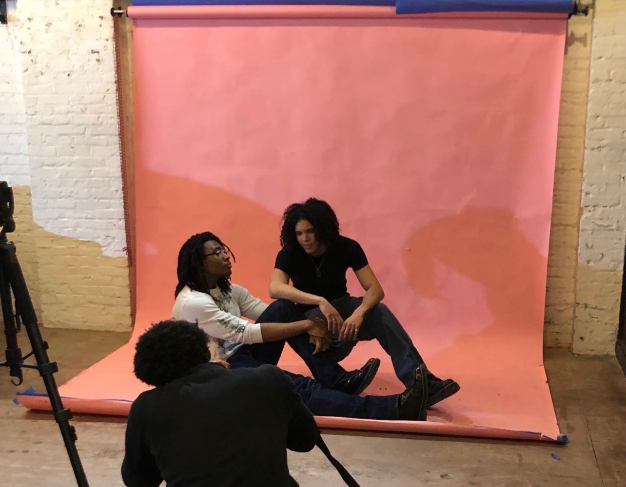 Two women sit on a pink backdrop with a person lying down between them, while a person in the foreground records the scene.