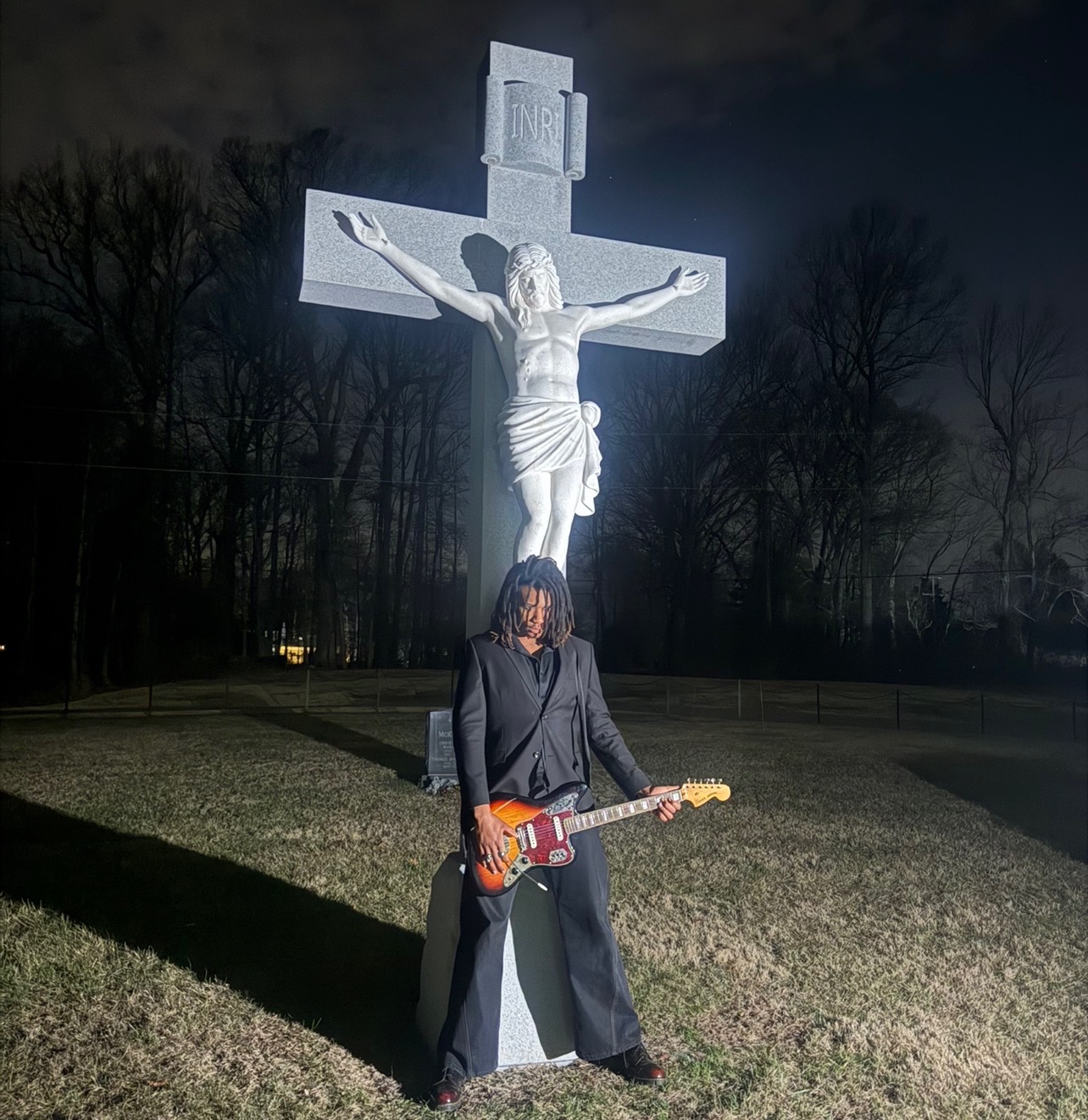 Person with long hair and dark clothing holding an electric guitar in front of a large crucifix statue outdoors at night.