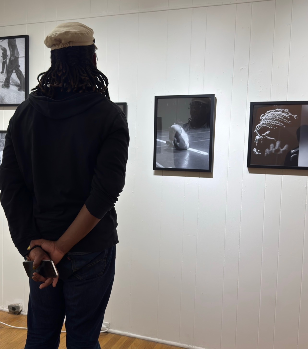 Person with a beige hat and dark clothing stands in front of framed photographs on a white wall, holding a phone behind their back.