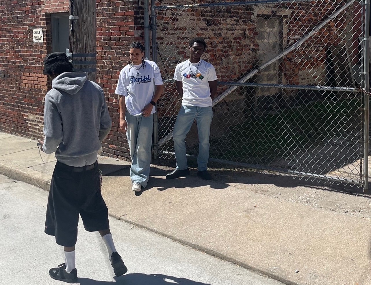 Three young men standing near a chain-link fence on a sidewalk, one facing away and two facing the camera, in casual clothing.