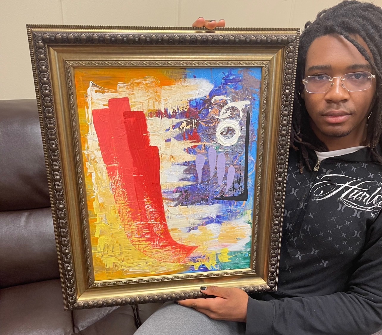 Person with glasses holding a colorful abstract painting in a decorative frame, sitting on a dark sofa.