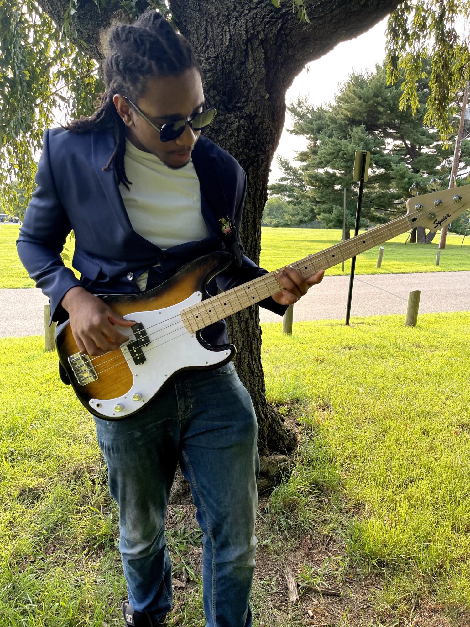 Person playing electric guitar outdoors near a tree, wearing sunglasses, a dark jacket, and jeans.