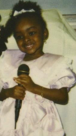 Young girl with a big smile holding a microphone, wearing a light-colored dress and a bow in her hair.