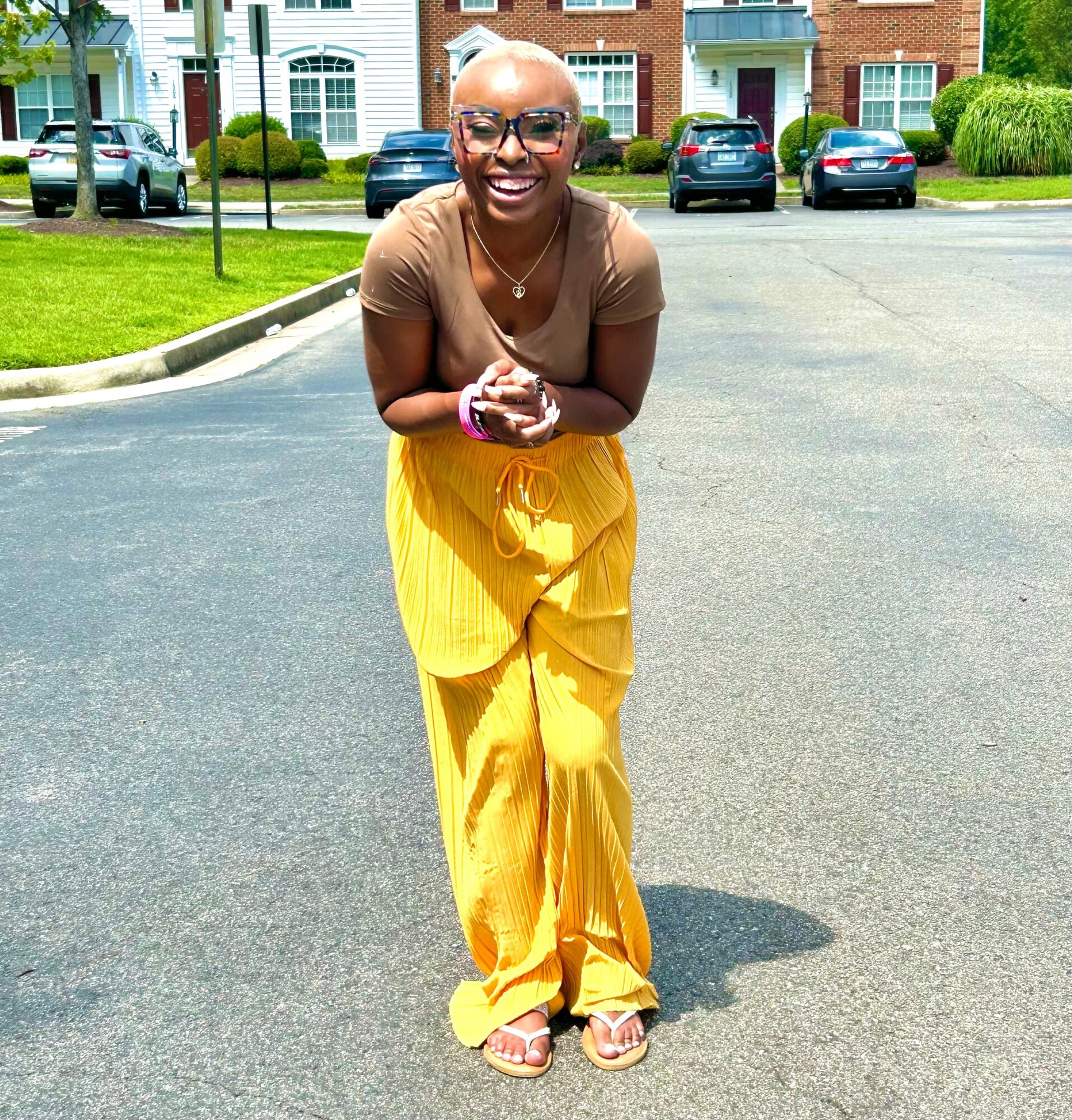 Smiling person with glasses and a shaved head, wearing a brown top and yellow pants, standing on a street.