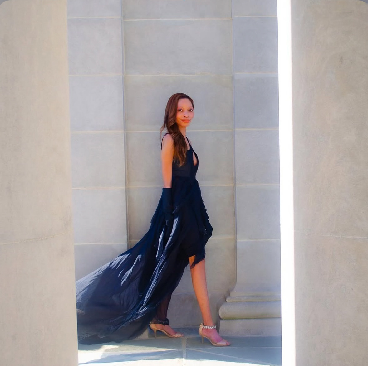 Woman in a dark dress and high heels standing between two large stone columns, with a flowing train behind her.