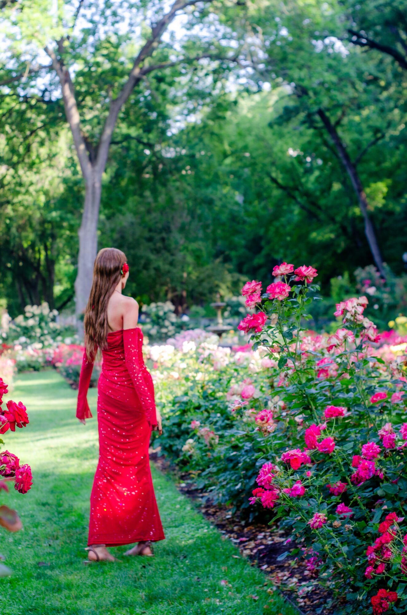 Girl in a red dress standing in a garden with pink and white flowers, surrounded by green trees.