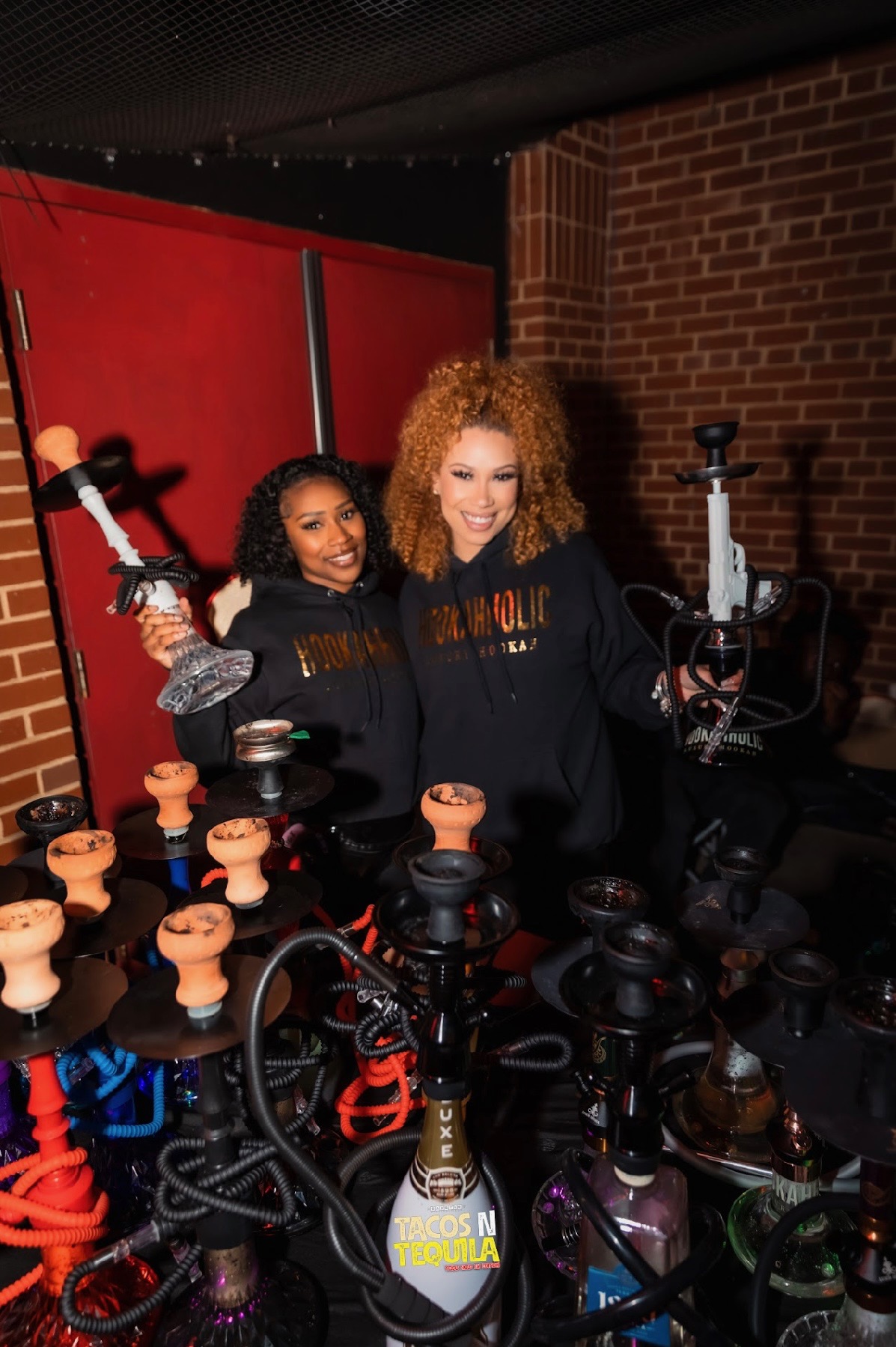 Two women smiling behind hookahs and a table with multiple hookahs, brick wall background, indoor setting.