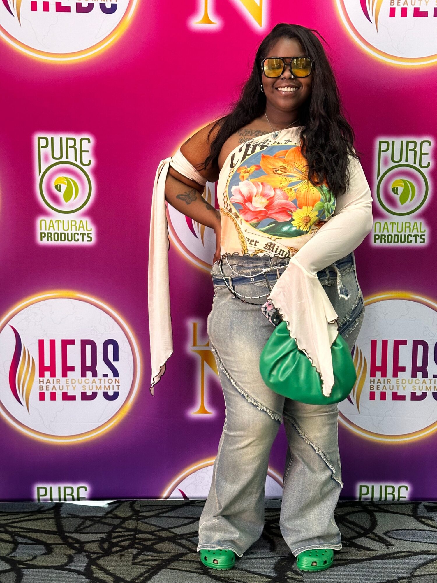 Woman with long dark hair wearing glasses, colorful top, light jeans, and green shoes, standing in front of a pink background with logos.