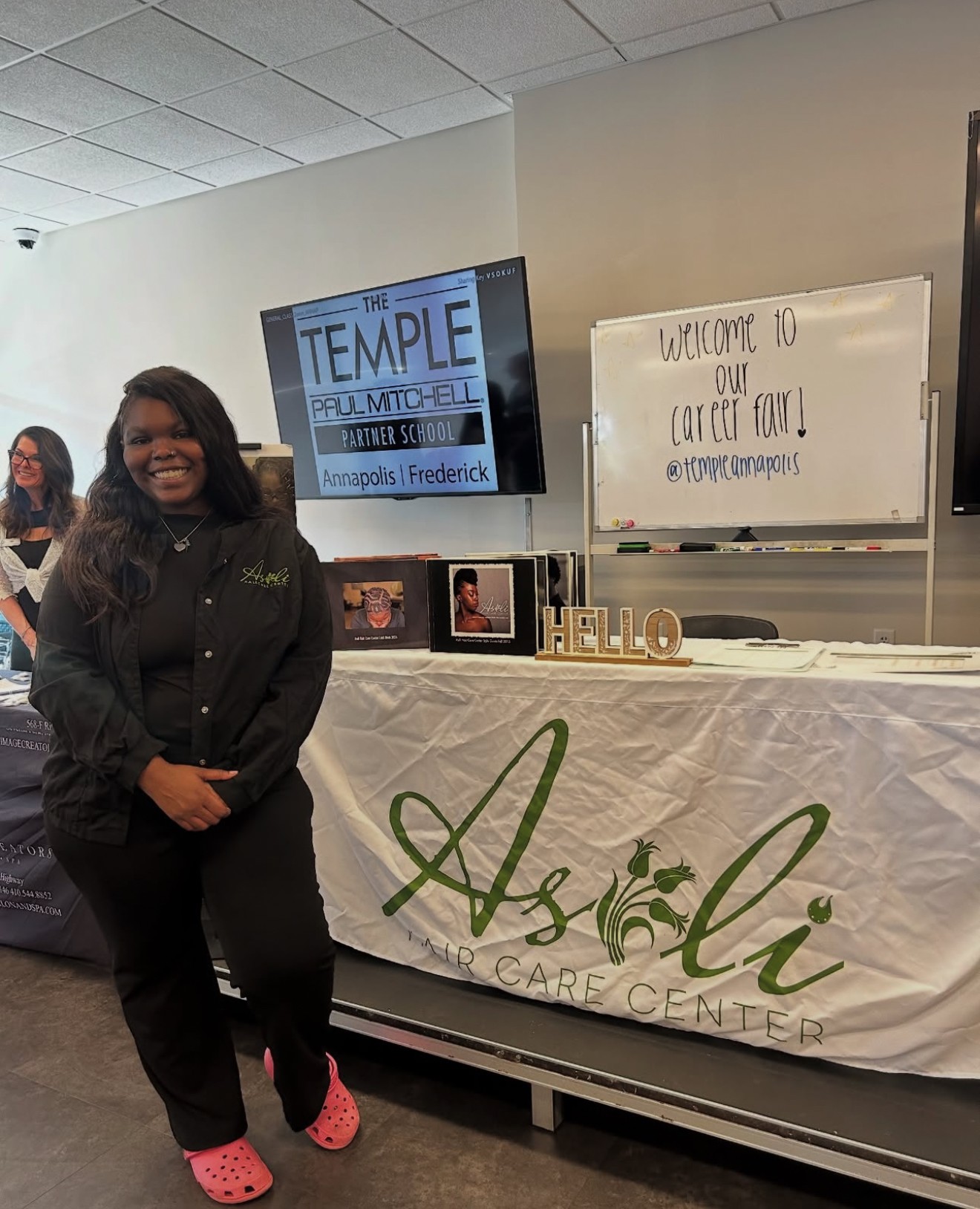 Woman standing in front of a table with a banner, whiteboard, and TV screen, smiling, indoors, casual attire, pink shoes.