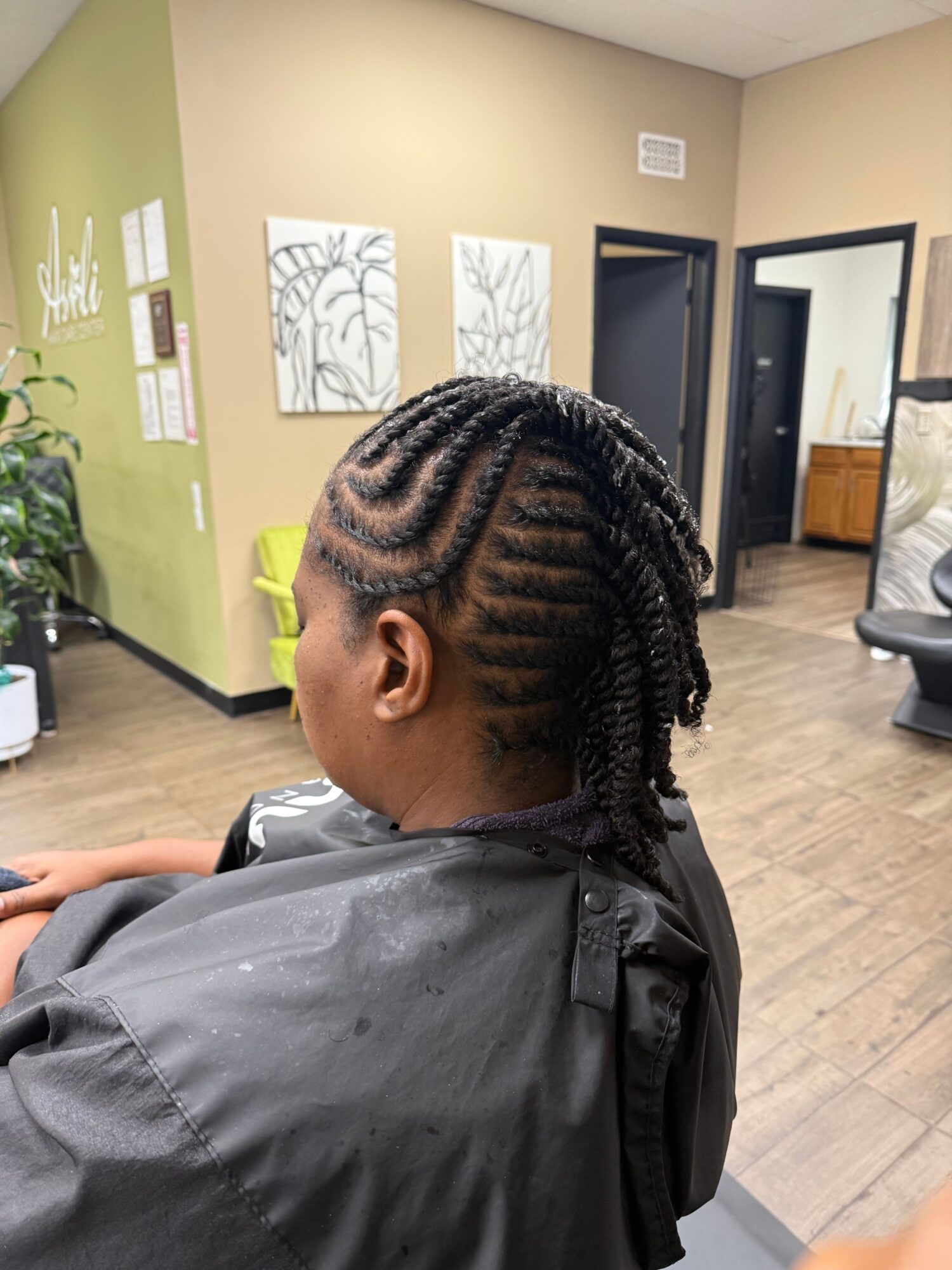 Person with braided hairstyle sitting in a salon chair, facing right, with salon interior in background.
