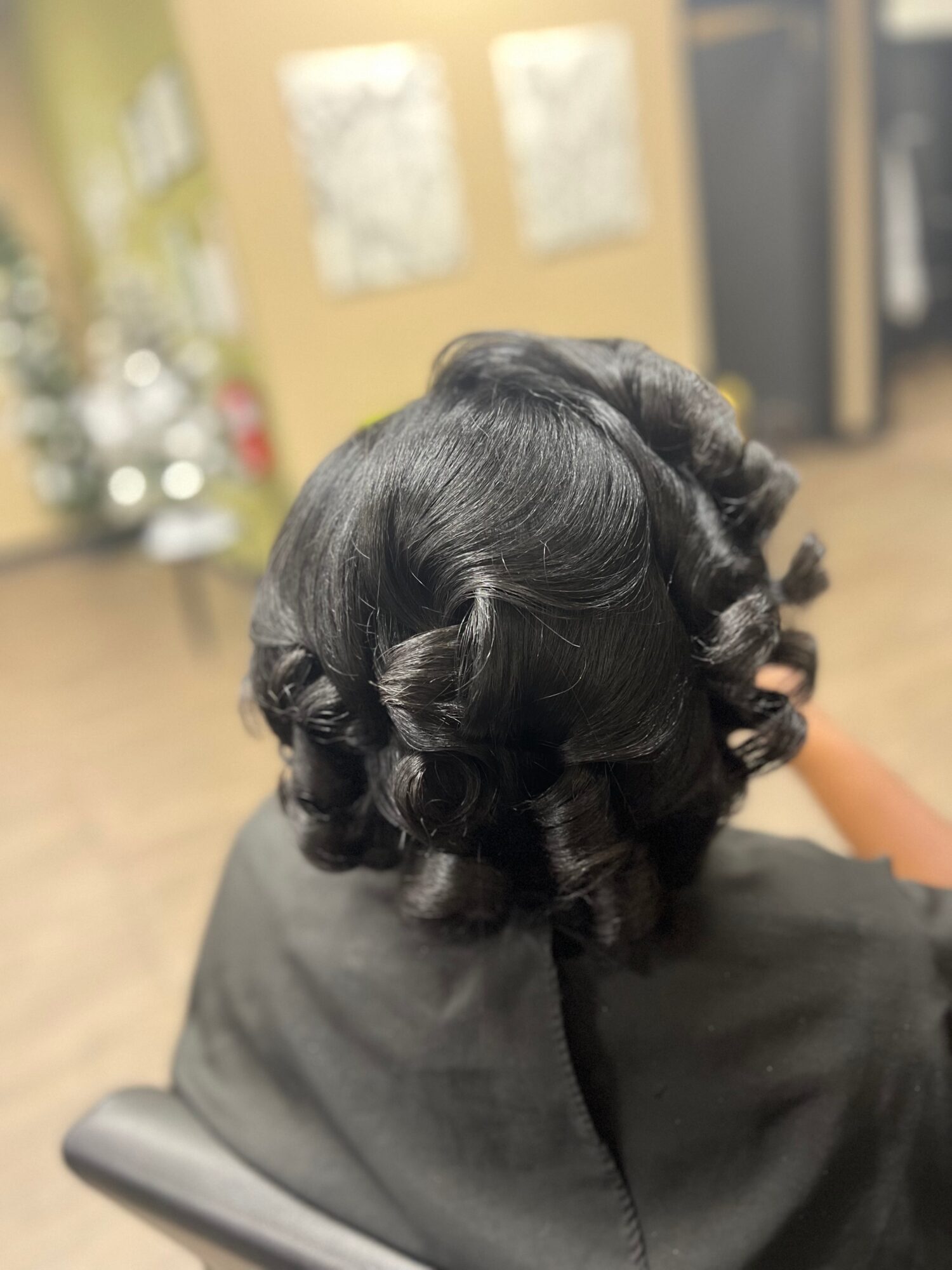 Person with styled black hair in curls, sitting in a salon chair, with a blurred background of salon interior.