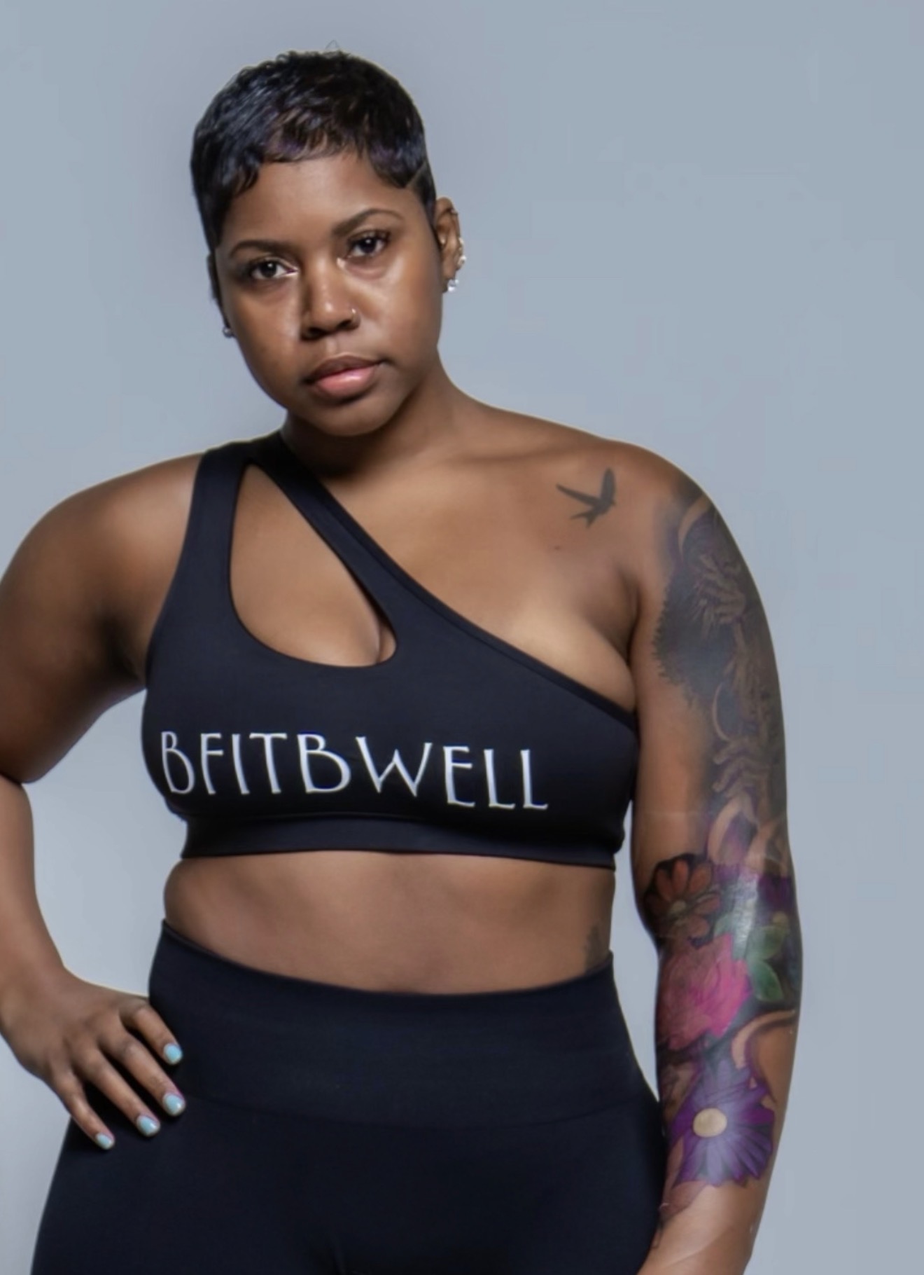 Woman with short hair wearing a black sports bra with 'BEITBWELL' text, standing with hand on hip, colorful tattoo on arm.