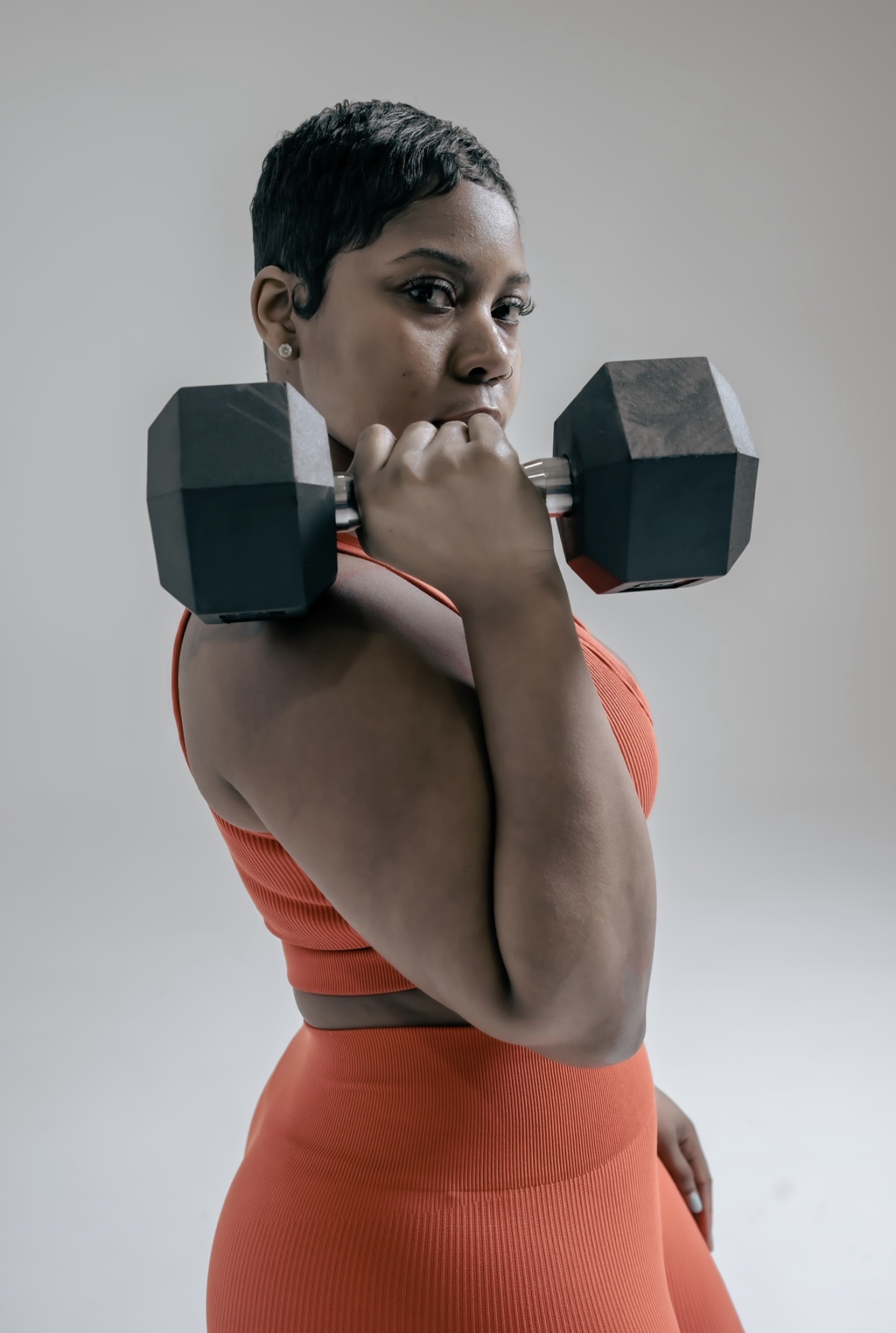 Woman with short hair lifting a dumbbell on her shoulder, wearing an orange sports top and matching pants.