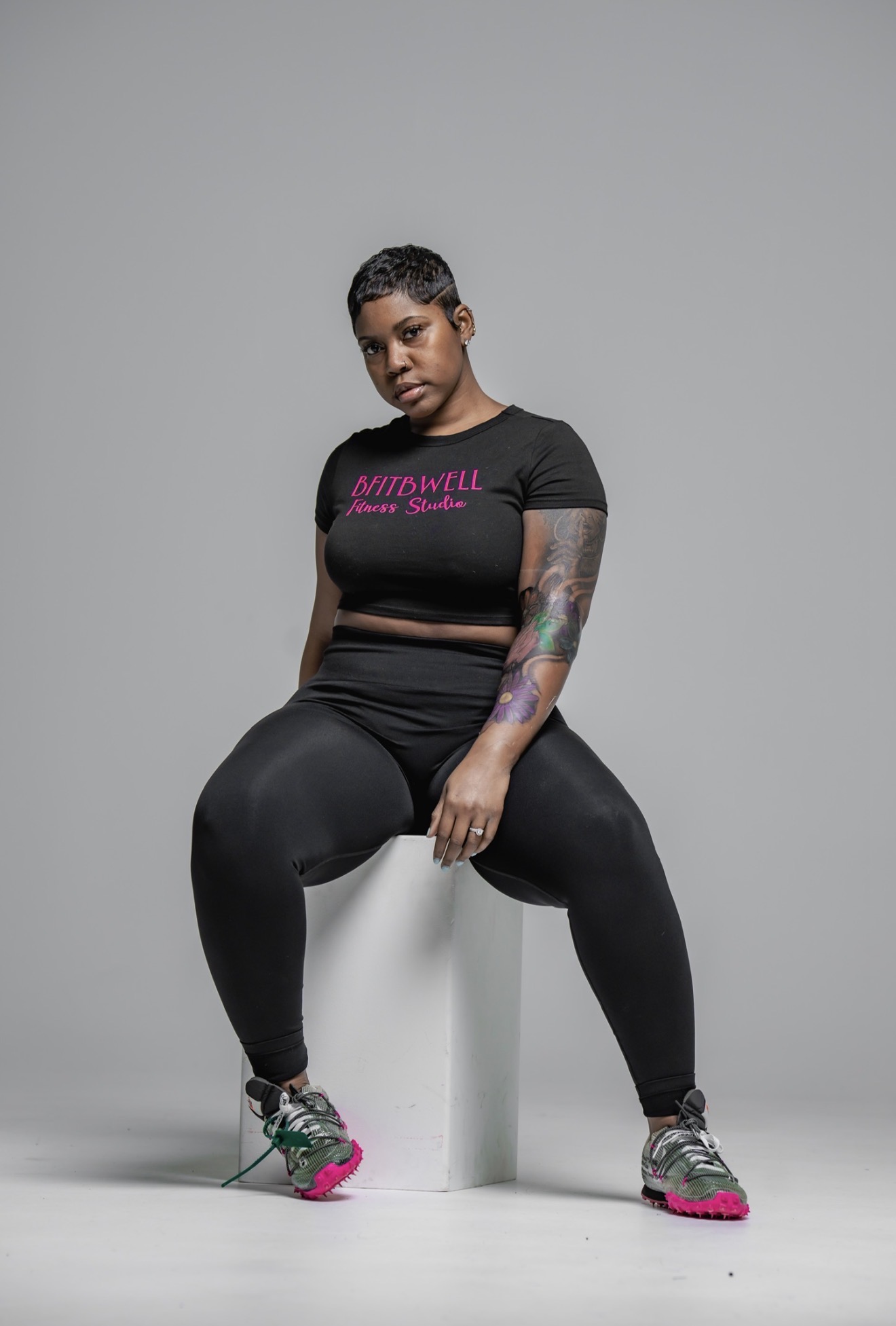 Woman with short hair and tattoos sitting on a white block, wearing a black t-shirt and black pants, against a gray background.