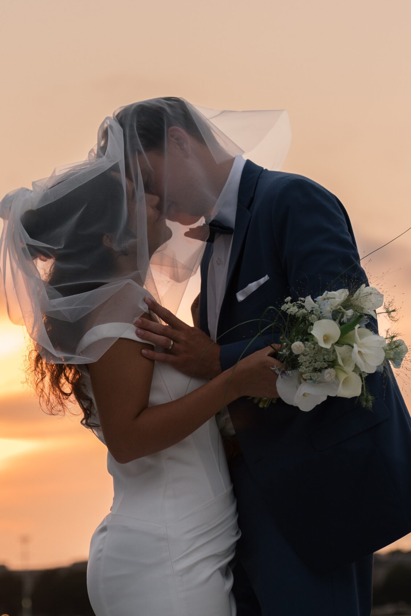 A bride and groom kiss outdoors at sunset, with the bride holding a bouquet of white flowers and greenery.