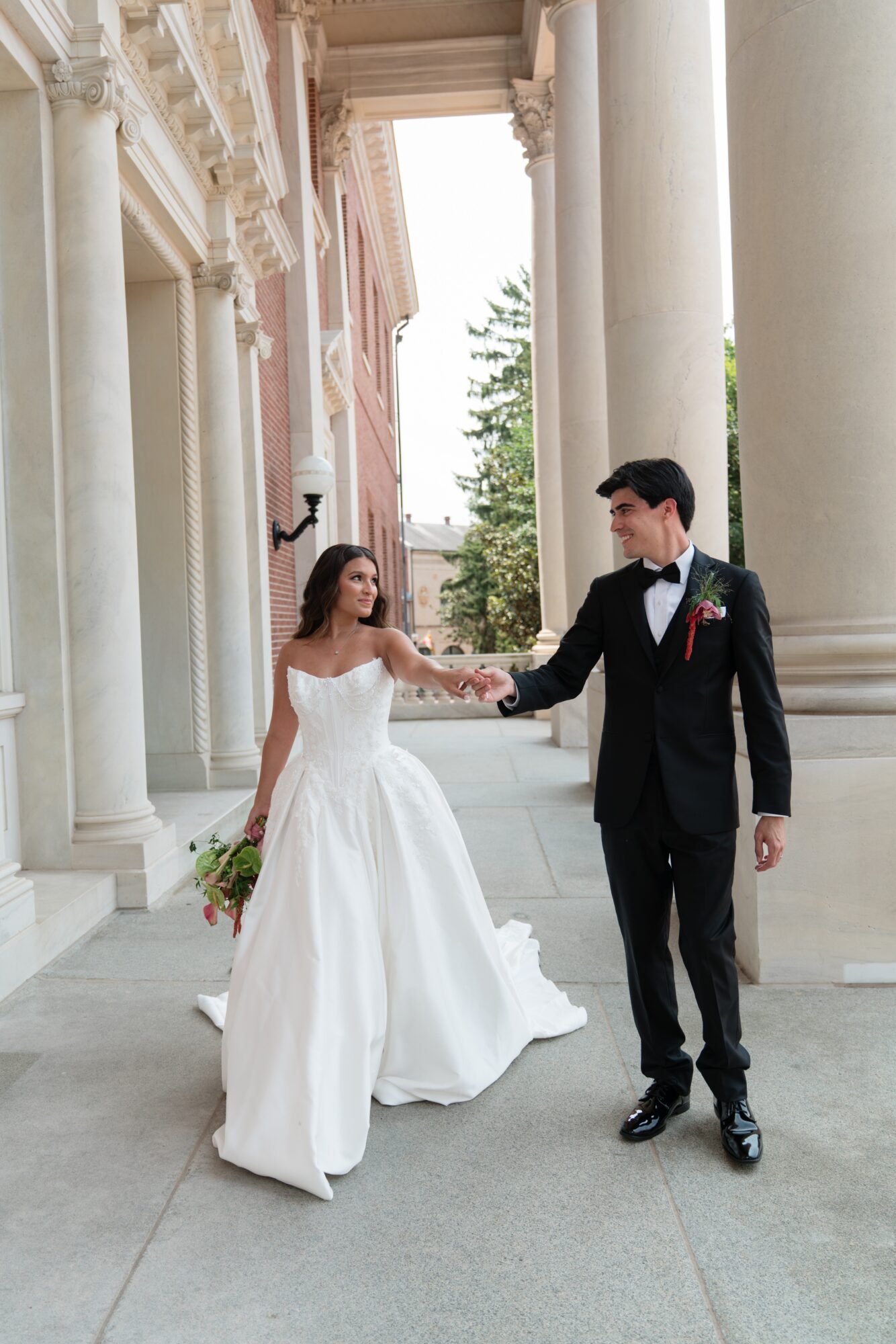 Bride and groom holding hands outside a grand building with large columns, bride in a white gown, groom in a black tuxedo.