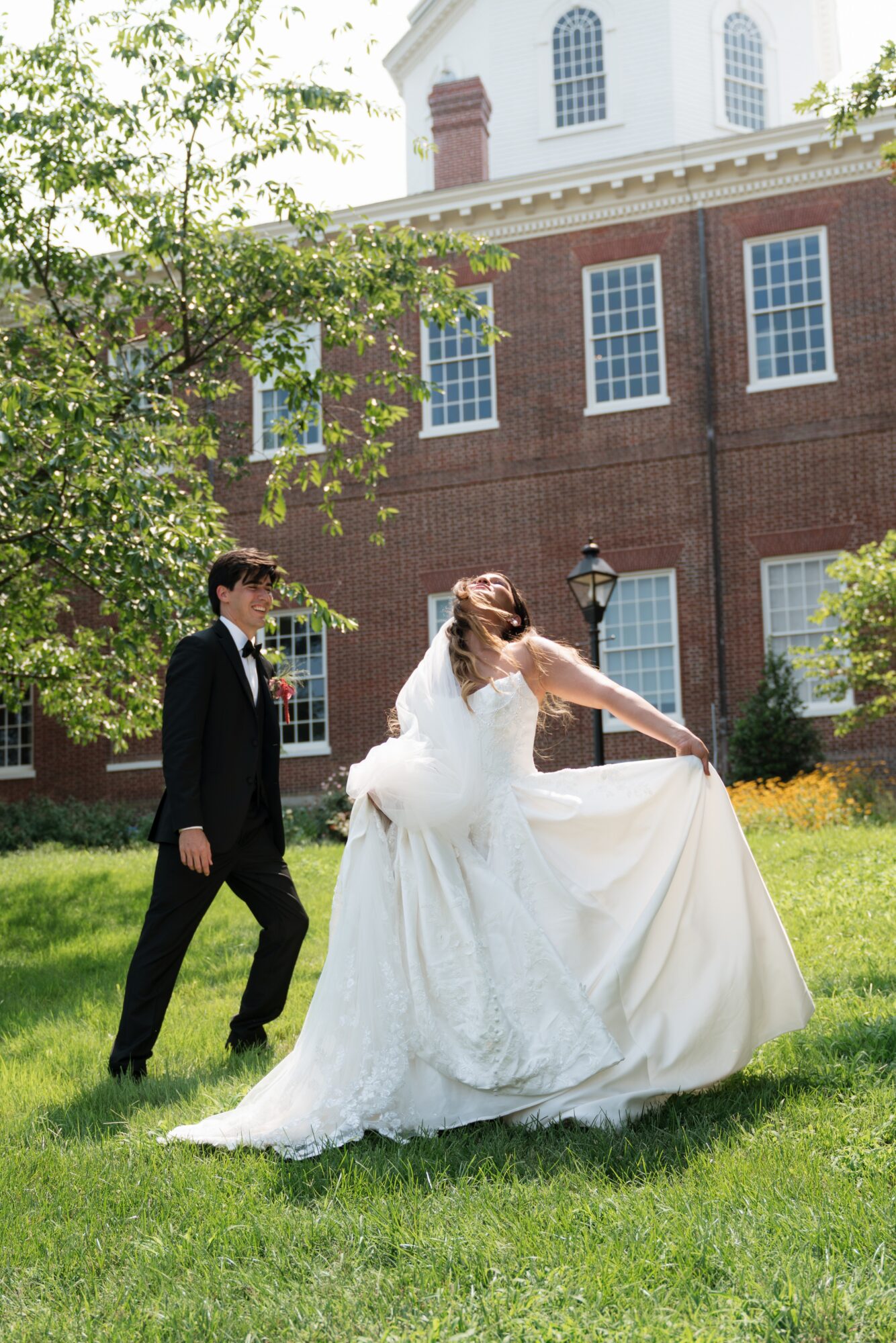 Bride in a white wedding dress twirling, groom in a black suit watching, green grass and a brick building in background.