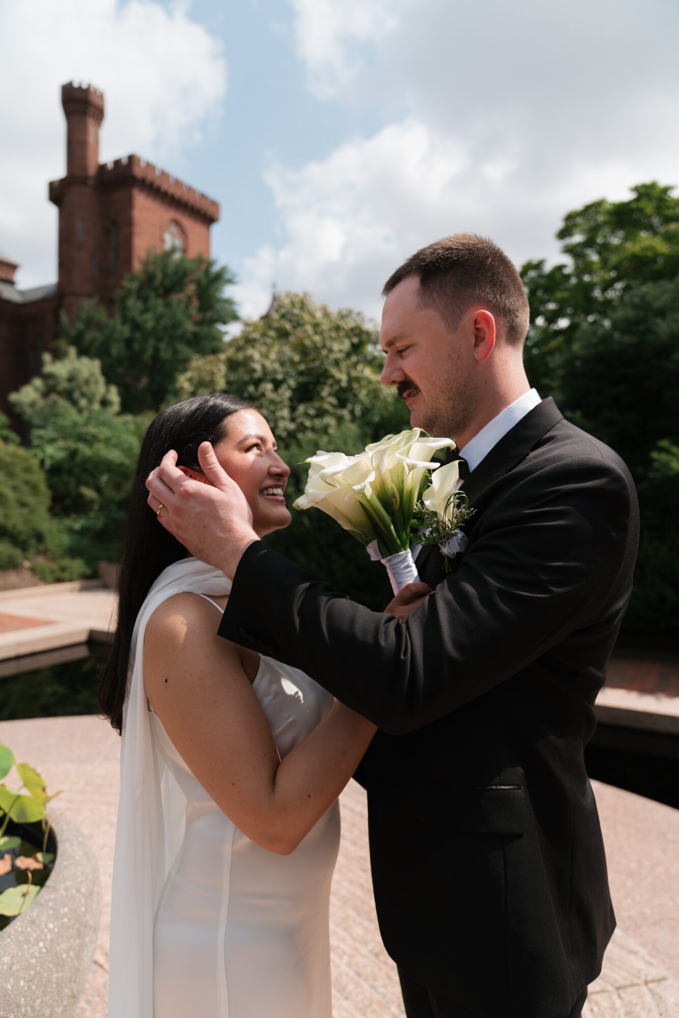 A couple in wedding attire smiling and holding flowers outdoors, with a building and trees in the background.