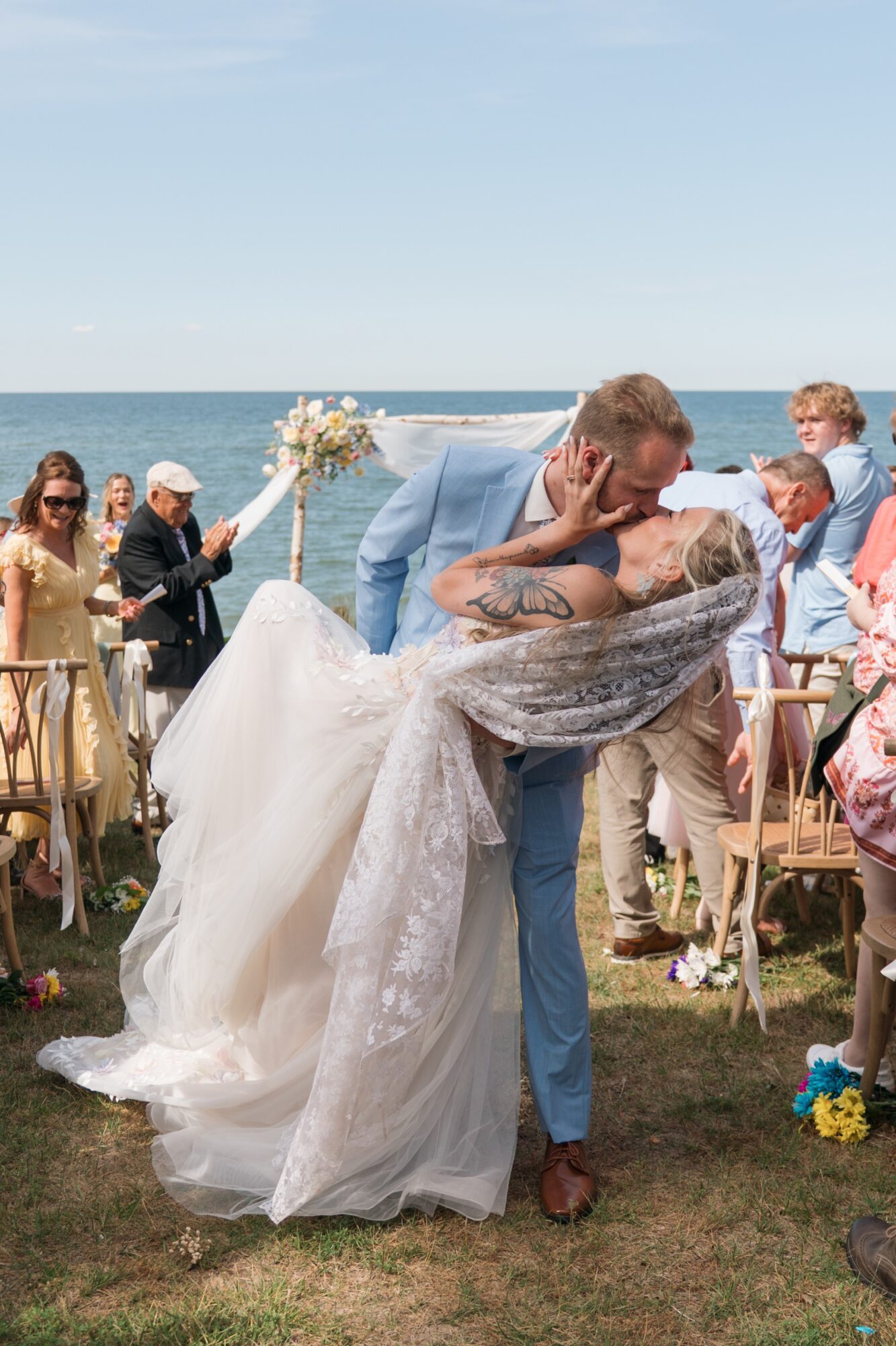 A couple shares a kiss outdoors during a wedding ceremony, with guests and a floral arch in the background.