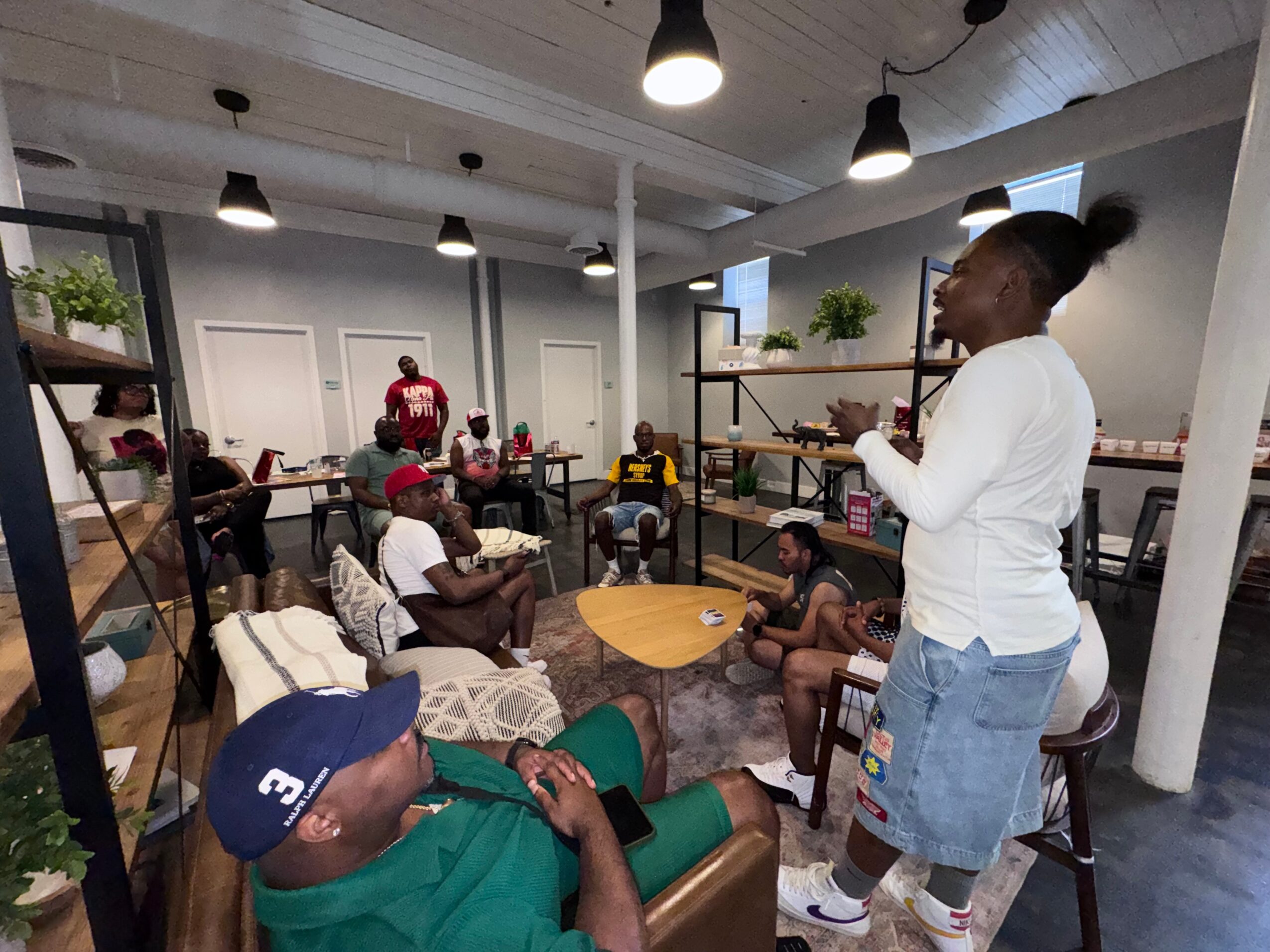 Group of people sitting and standing in a modern room with white walls, plants, and hanging lights, engaging in conversation.