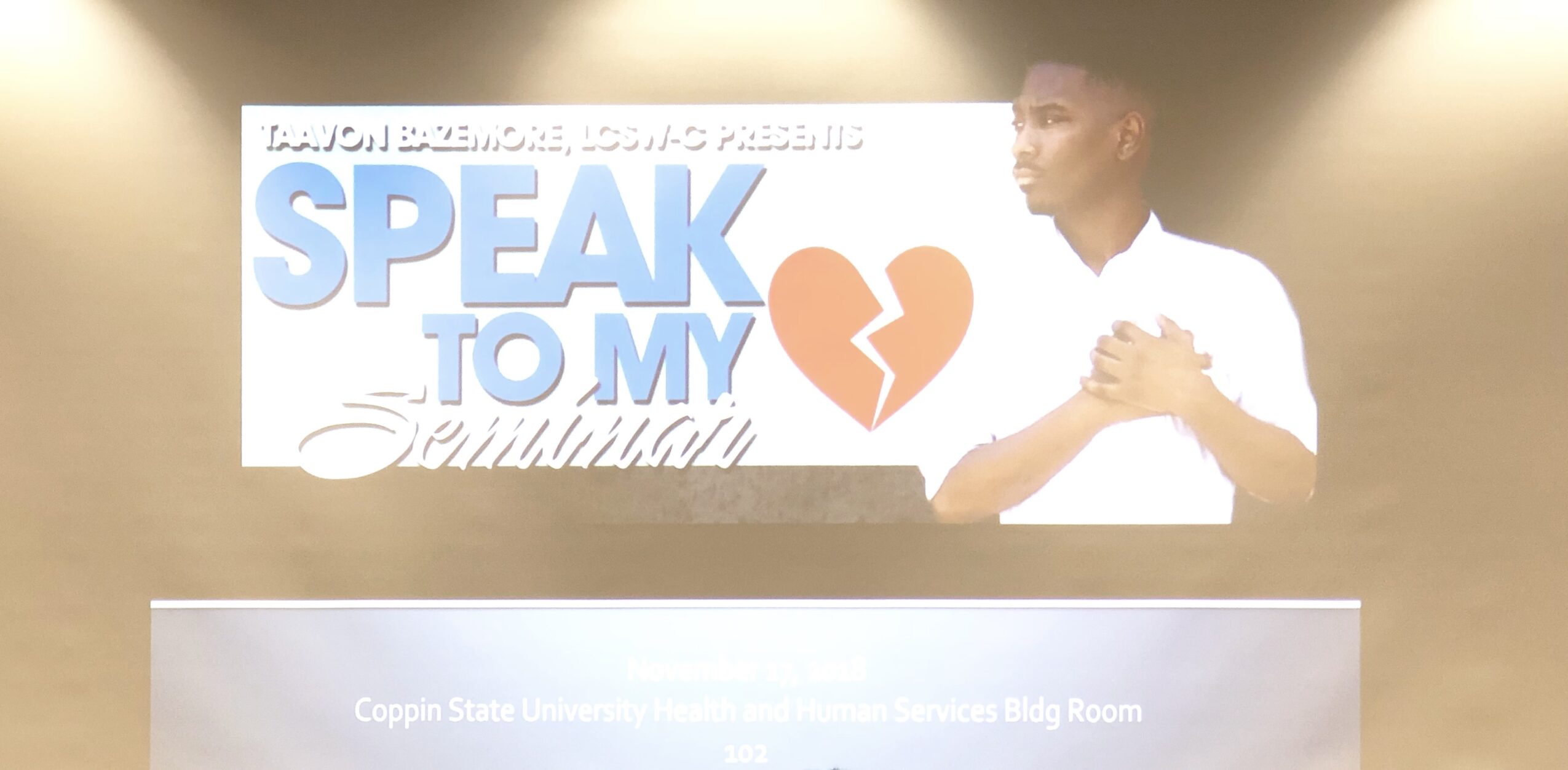 A person standing in front of a screen with text and a broken heart symbol, holding hands over chest, in a well-lit room.