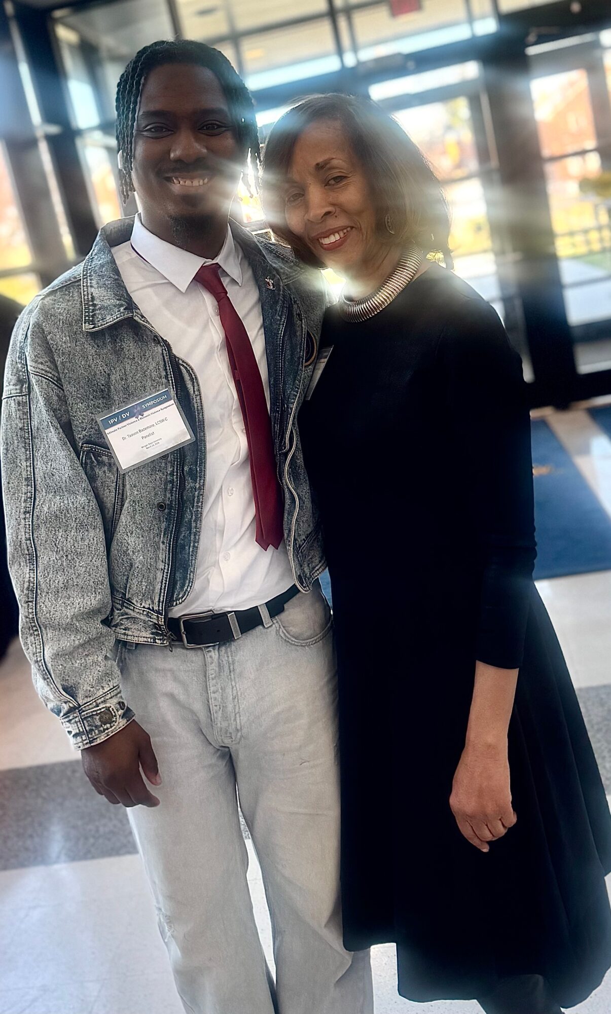 Two people standing together indoors, smiling, with sunlight and windows in the background.