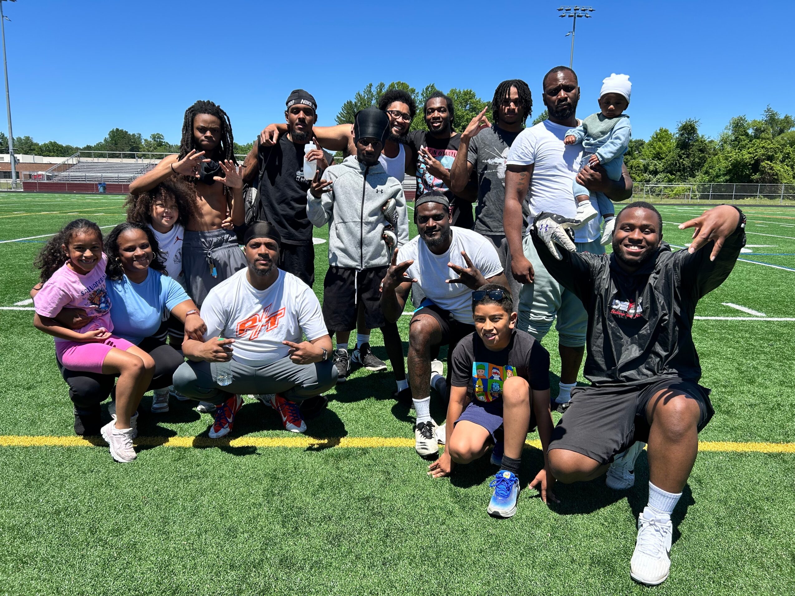 Group of people, including children, posing on a sports field under a blue sky, some making gestures, smiling.