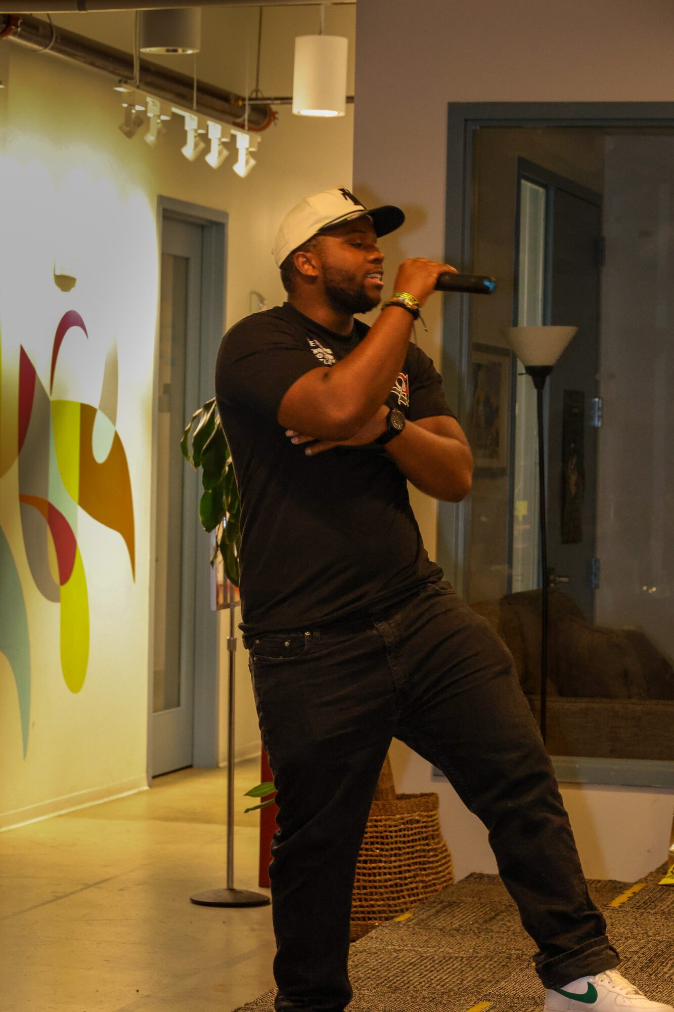 Man wearing a white cap and black shirt speaking into a microphone indoors, with a window and decorative wall behind him.