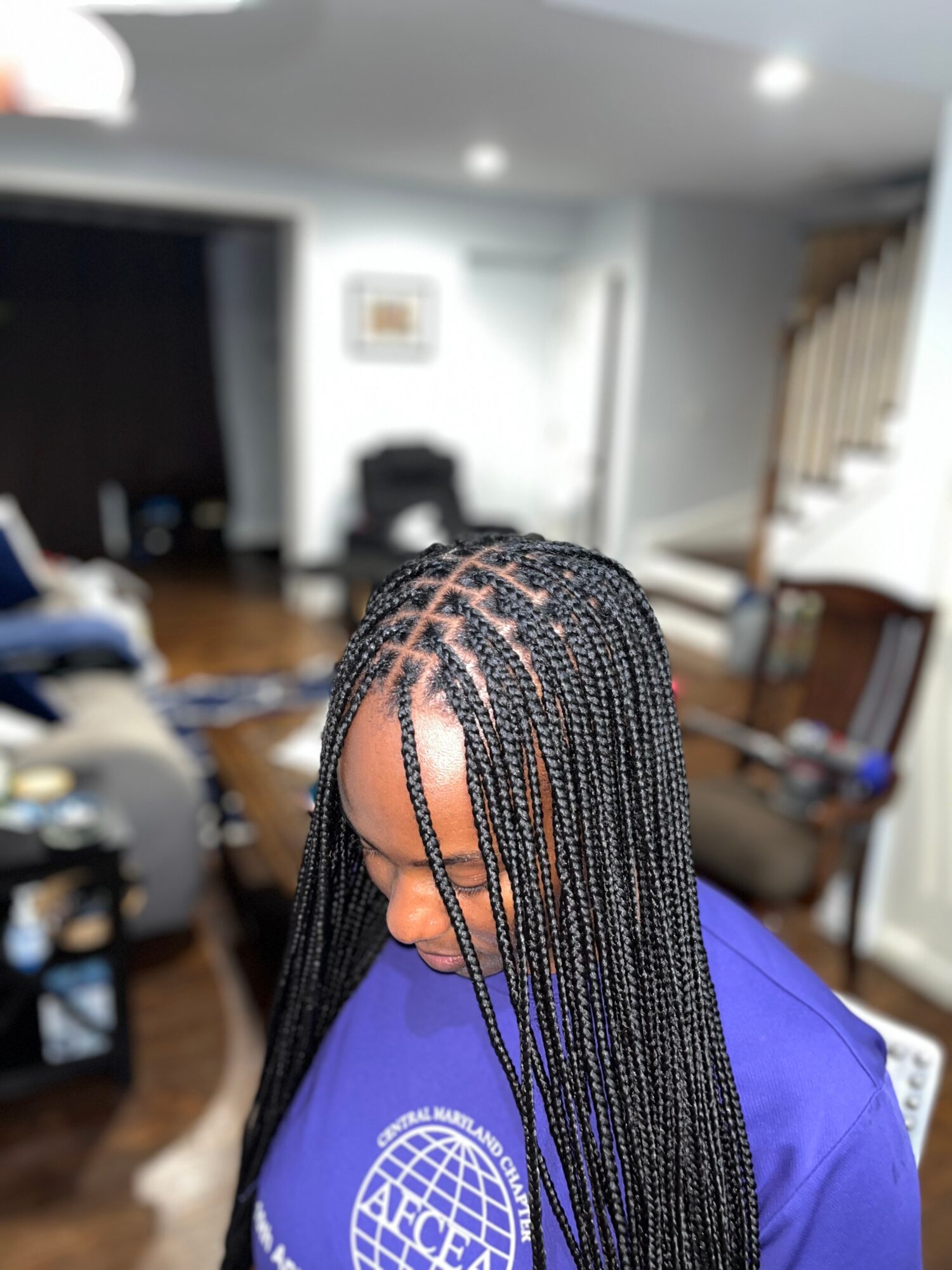 Person with braided hair wearing a purple shirt, indoors with furniture and stairs in background.