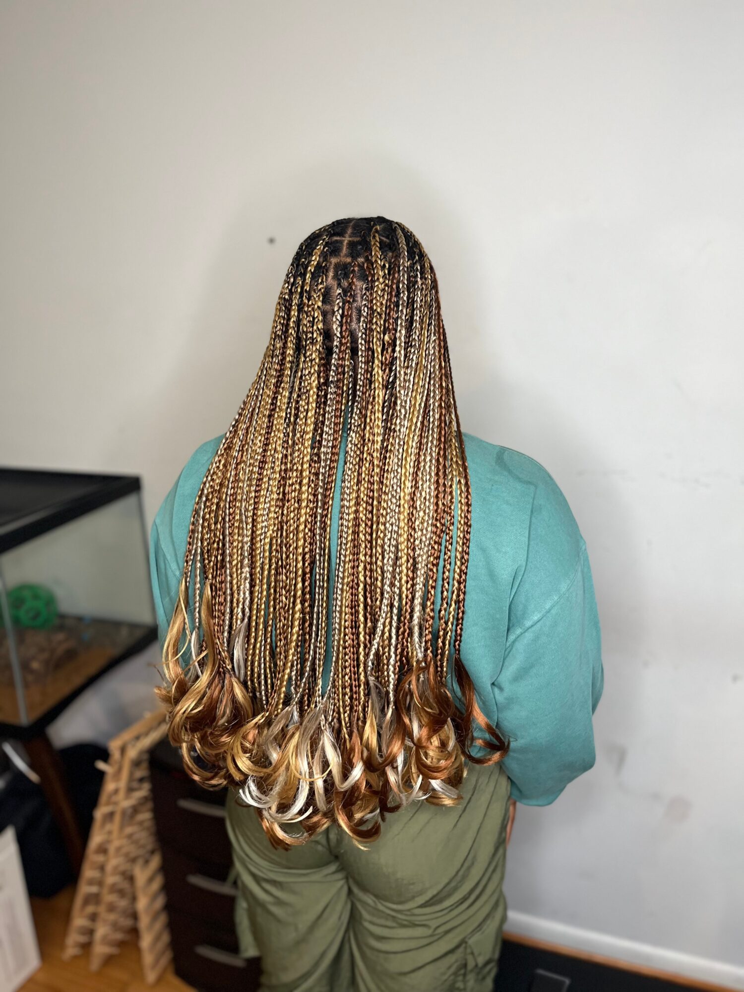 Person with long braided hair standing with back to camera, wearing a teal top and green pants, against a plain white wall.