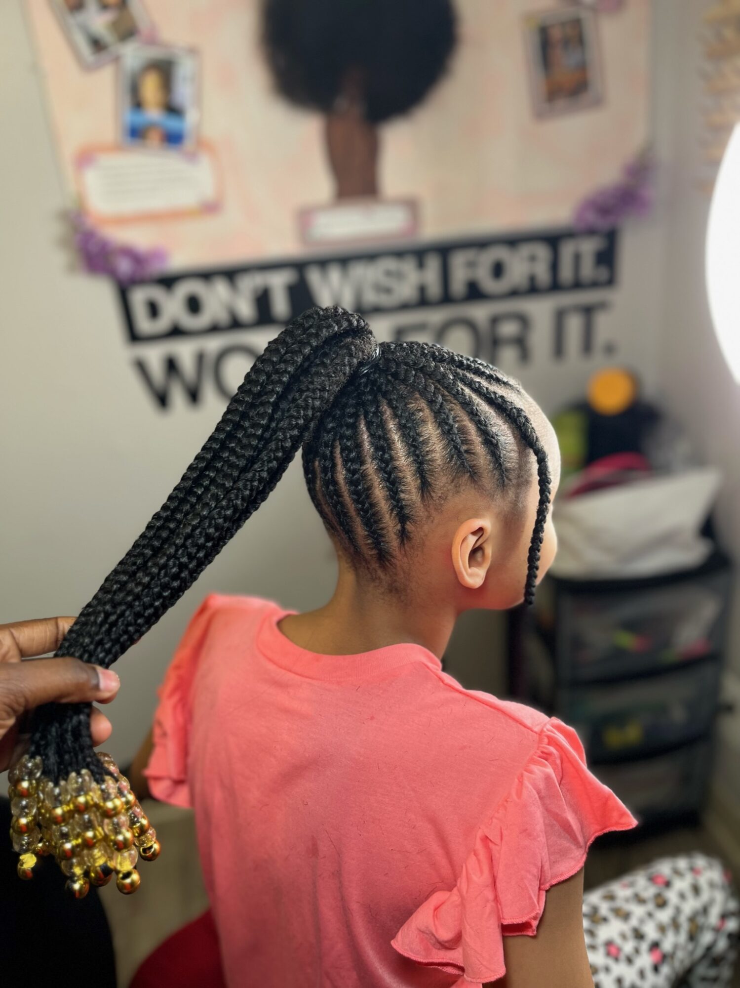 Child with braided hairstyle, wearing a pink top, facing away, with a poster on the wall behind.