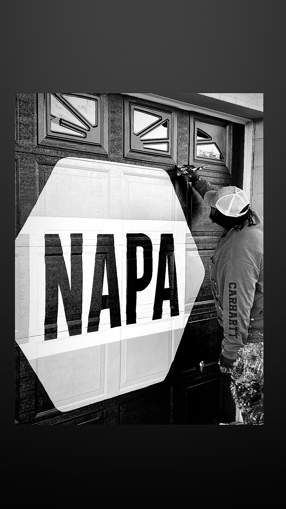 Person painting a large NAPA sign on a wall near windows, wearing a cap and patterned pants, in black and white.