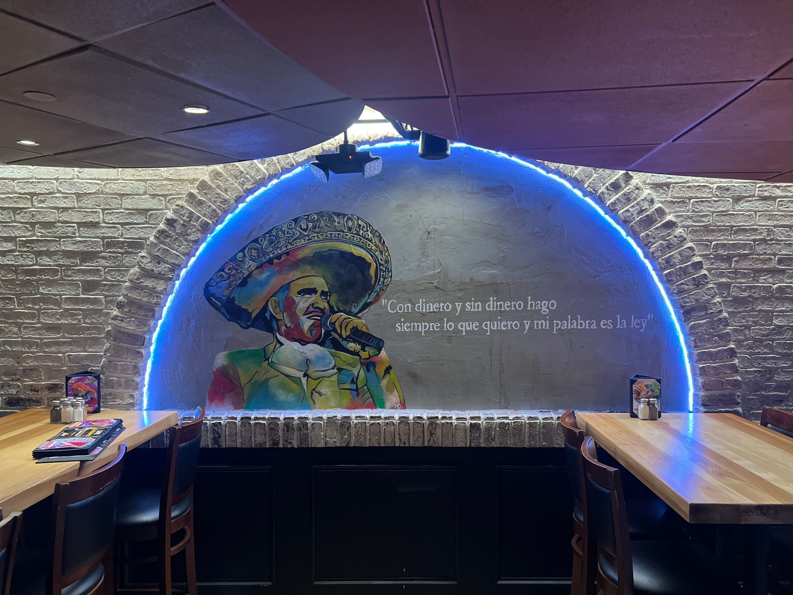Wall mural of a man in a sombrero with a microphone, illuminated by blue neon lights, with tables and chairs in front.
