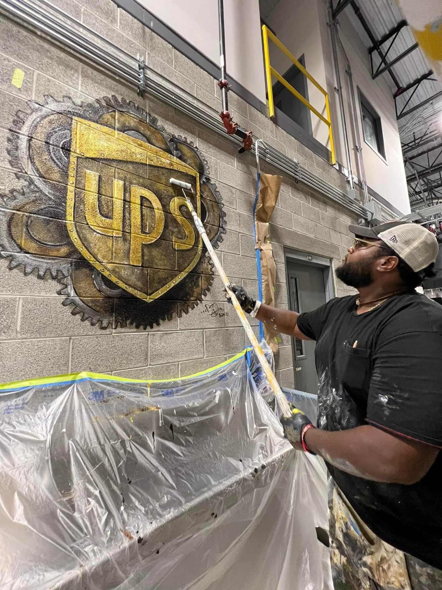 Man spray-painting UPS logo on wall in industrial setting, wearing cap and glasses, with plastic covering nearby surfaces.
