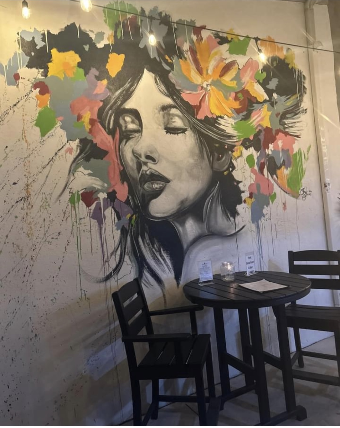 Wall mural of a woman's face with colorful floral and abstract elements around her head, in a room with tables and chairs.