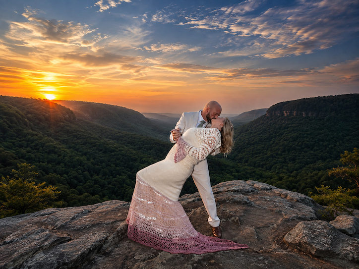 A couple kissing on a rocky ledge during sunset with a valley and sky in the background.