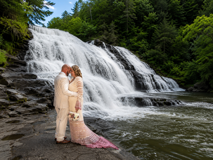 Couple standing close by a waterfall surrounded by trees, with water flowing over rocks and lush greenery in the background.