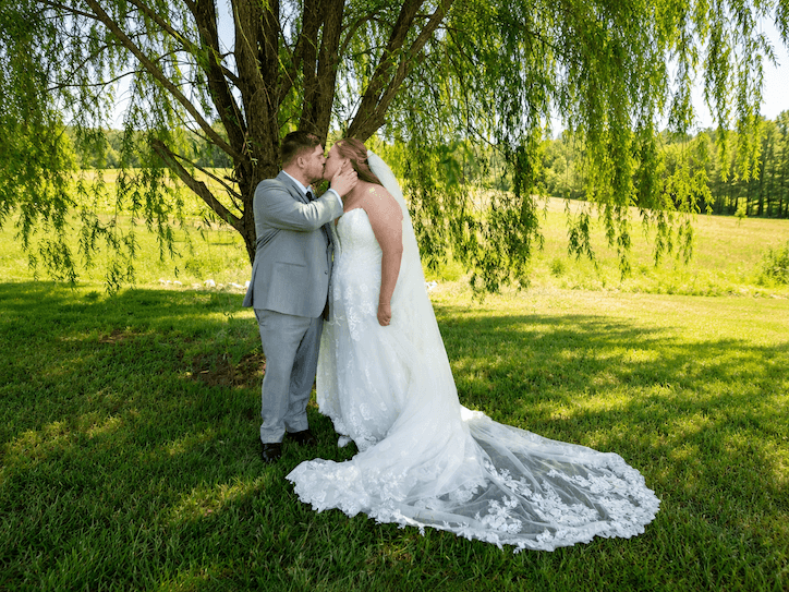 A couple in wedding attire stands under a tree, kissing outdoors on a sunny day, with a grassy field in the background.