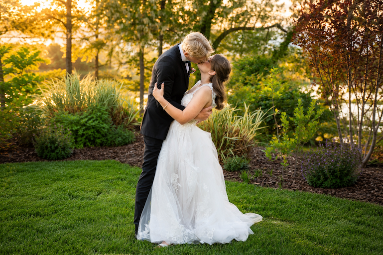 Couple in wedding attire sharing a kiss outdoors with trees and greenery in the background.