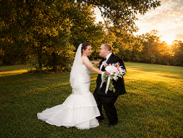 A bride and groom stand outdoors on a grassy field with trees and sunset in the background, holding hands and facing each other.